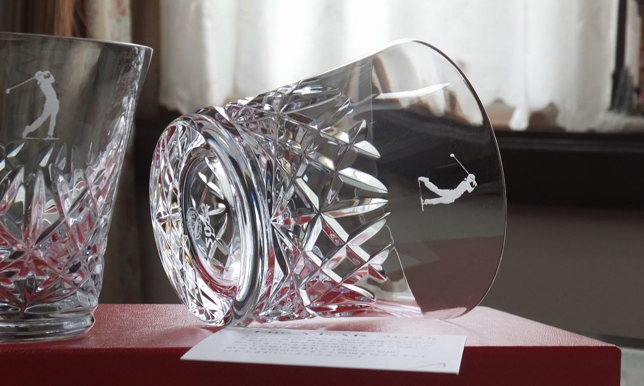 バカラのアリア（Aria）タンブラー2025のクリスタルグラスのクローズアップ写真。グラスの側面中央に、ゴルフ記念品として『スイングをしているゴルフプレーヤーのシルエット』が名入れ彫刻されているのが鮮明に確認できる。しずくを思わせる繊細なカットが特徴のタンブラーの透明度の高い面に、躍動感あふれるゴルフシーンが刻まれ、ゴルフコンペの優勝や特別なホールインワンを記念する世界最高峰の輝きを持つ記念品。