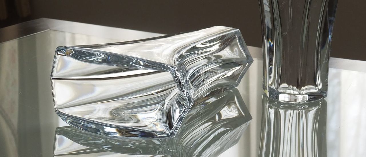 バカラの『ギンコ ベース（花瓶）』が、モダンなテーブルの上に横に倒して置かれている様子の写真。イチョウの葉をモチーフにした優美で流れるような曲線を持つベースの形状が、横向きに配置されることでより強調されている。クリスタルの透明感と、テーブルの表面との光のコントラストが際立ち、アートピースとしての洗練されたデザインと高級感を視覚的に伝えている。