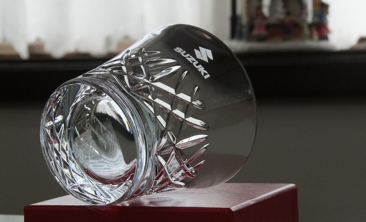 バカラの『アリア タンブラー 2025（イヤータンブラー）』の側面に名入れ彫刻を施した退職祝い記念品の制作実績。空気が振動するさまをイメージしたデザインのタンブラーの側面に、スズキ自動車のロゴマークが精巧に刻印されている。年号入りの限定品に、長年にわたり貢献した企業のシンボルマークを刻むことで、長年の功績に対する深い敬意と、格式ある企業記念品としての特別な価値を視覚的に伝えている。