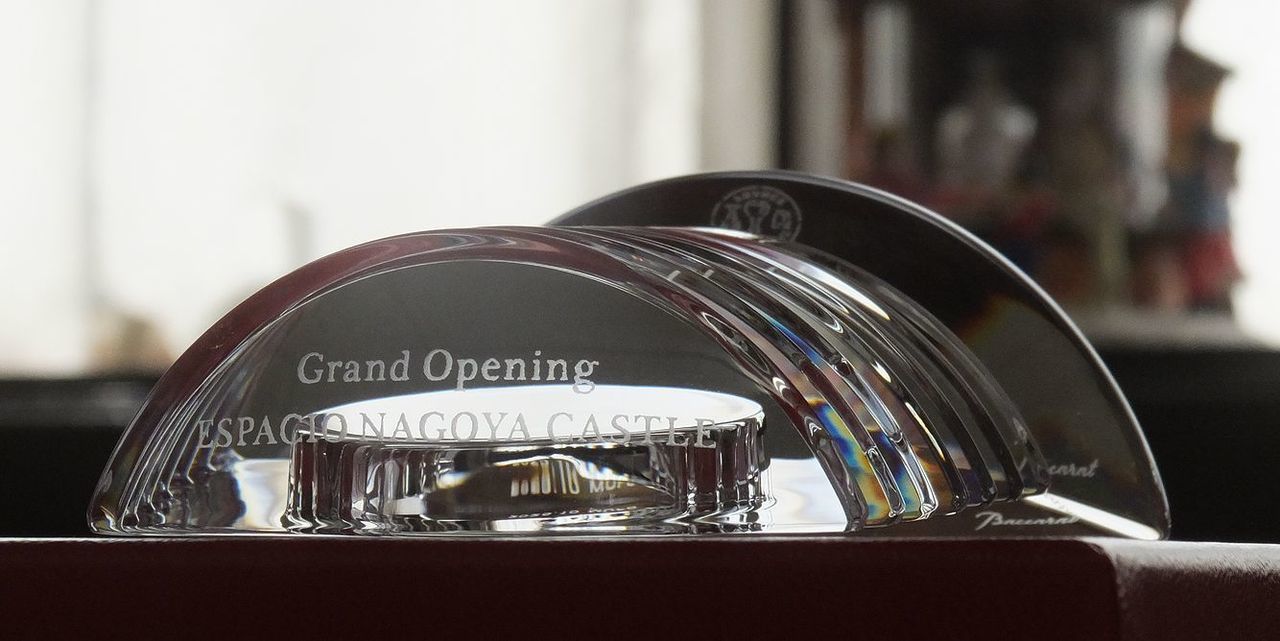 バカラの置き時計、ラランドクロック（Lalande Clock）のクリスタル製クロックのクローズアップ写真。アール・デコ調の優美なフォルムと、繊細なダイヤモンドカットが特徴的な時計の上部（カーブしたクリスタル部分）に、『Grand Opening ESPACIO NAGOYA CASTLE』という開店・開業記念のメッセージが名入れ彫刻されている。時間の正確さと不変のクリスタルの輝きが、新たな事業の成功と門出を祝う格式高い記念品