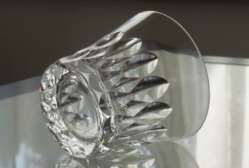 バカラのイヤーグラス、ダリア（Dahlia）タンブラー2026のクリスタルグラスが、テーブルの上に横に倒して置かれている写真。ダリアの花弁をモチーフとしたダイナミックかつ複雑なV字カットが、長い側面を通して光を反射させ、グラスの優美なフォルムを際立たせている。重厚な底面と、繊細なカットが描く光の軌跡が、クリスタルガラスの最高峰としての存在感と芸術的な美しさを強調した情景。