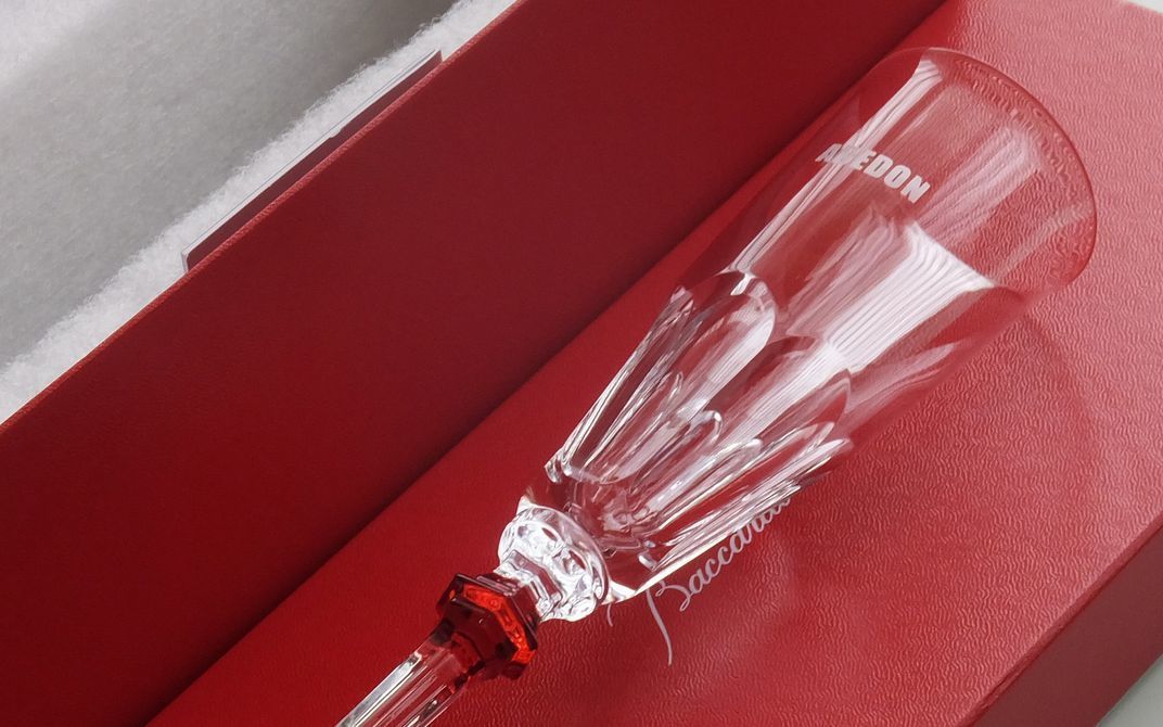 バカラ（Baccarat）の『アルクール イヴ シャンパンフルート』の側面に、アーティストオリジナルのロゴマークが精緻に名入れ彫刻された特別ギフトの写真。アイコンであるアルクールの重厚なフラットカットをエレガントでスレンダーなフォルムへと昇華させた最高級クリスタルグラスに、世界観やアイデンティティを象徴する刻印が美しく映え、ライブ・公演の開催記念やアニバーサリー、アーティストへの敬意と祝福を込めて贈る最高峰のメモリアルギフトイメージ。