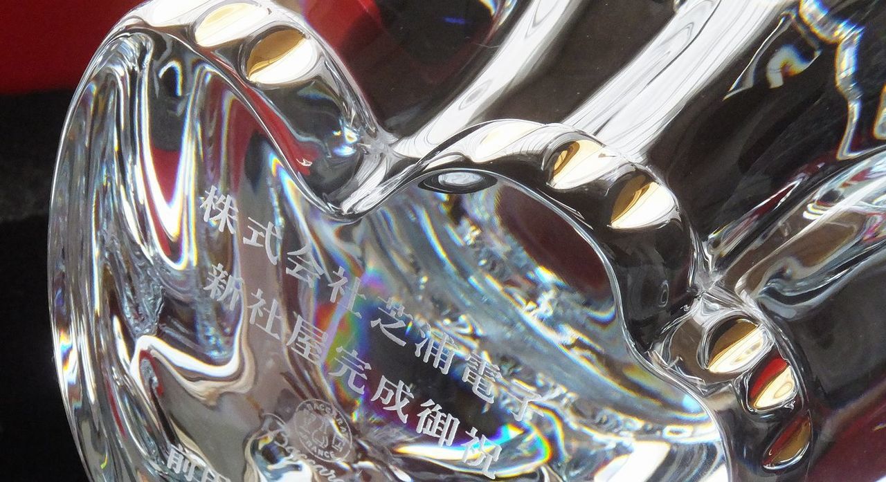 バカラ（Baccarat）の『まねき猫 XL』の底面に、『株式会社芝浦電子 新社屋完成御祝』と『企業名』の文字が精緻に名入れ彫刻された写真。圧倒的な存在感と輝きを放つ大型クリスタルガラスの招き猫に、新社屋の完成と更なる事業発展を祝う特別な刻印が美しく施され、商売繁盛と千客万来を願う最高峰の竣工記念品や企業間のお祝いギフトにふさわしい、重厚感と気品を備えた格調高いイメージ