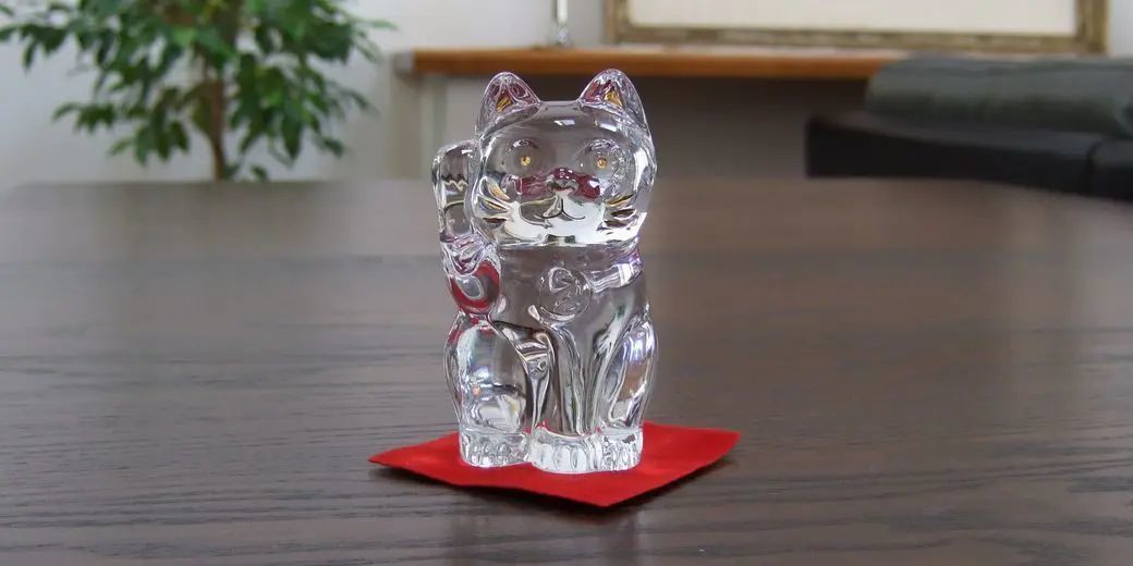 バカラの招き猫 クリアが、テーブルの上に置かれている写真。透明なクリスタルでできた、愛らしい招き猫が、上品な輝きを放っています。幸運を招くとされる招き猫は、開店祝いや開業祝い、または事業の発展を願う贈り物として、親しみやすくも格式ある法人ギフトとして喜ばれる逸品です。