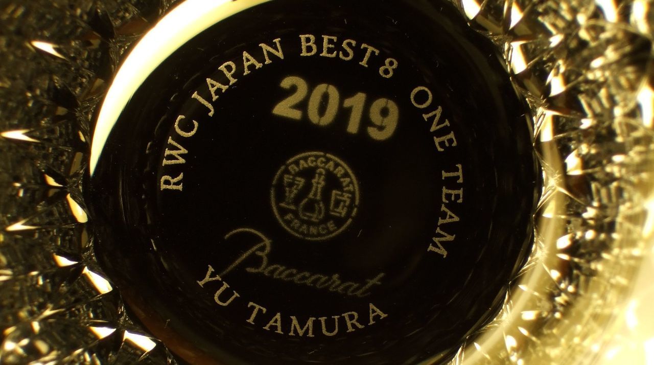 バカラの2019年限定イヤーグラス『ヴィータ』の底面に、ラグビーワールドカップ2019日本大会のベスト8進出を祝した名入れ彫刻を施したサンプル画像。グラスを覗き込んだ際に見える底面に、『RWC JAPAN BEST8』『ONE TEAM』の文字と『受贈者のお名前』が、力強く結束を感じさせる書体で精緻に刻印されている。グラス側面の複雑なカットが光を反射し、日本中が熱狂した歴史的快挙とチームの絆を象徴しており、一生に一度の感動を最高級のクリスタルに封じ込めた、誇り高き特別なアニバーサリーギフトであることを視覚的に表現している。