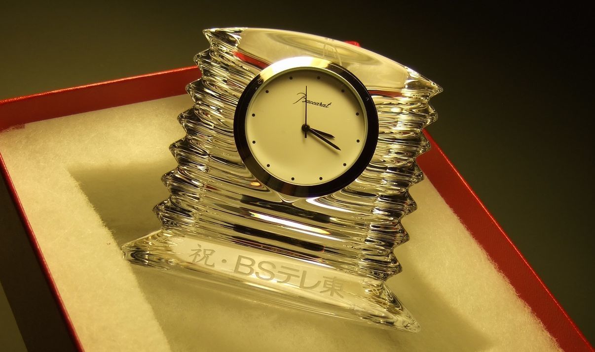 バカラのクリスタル製置き時計『ラランドクロック』に、社名変更を祝した名入れ彫刻を施した記念品のサンプル画像。透明度の高い円形クリスタルの下部に、『祝・BSテレ東』の文字が、お祝いにふさわしい華やかで力強い書体で精巧に刻印されている。天文学者の名を冠したクロックが放つ「星の輝き」のような光の反射が、新しい社名でのさらなる飛躍と輝かしい未来を象徴しており、企業の重要な節目を祝う格調高いギフトであることを視覚的に表現している。